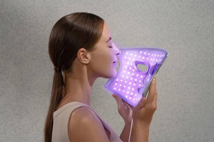 A person holds a glowing purple LED light therapy face mask against their face with eyes closed, against a neutral grey background.‍