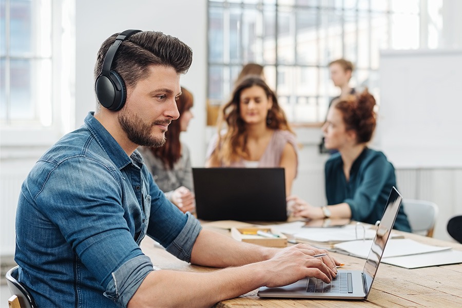 A man wearing noise-cancelling headphones, focused on his laptop in a busy office with colleagues collaborating around him.