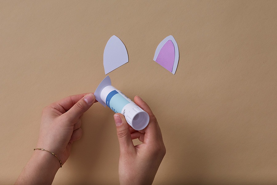 Hands using a glue stick to assemble paper unicorn ears, with one ear already completed with a purple inner section.