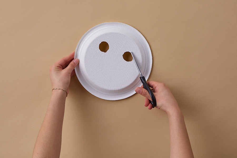 Hands cutting eye holes into a white paper plate to make a DIY animal mask on a plain brown background.