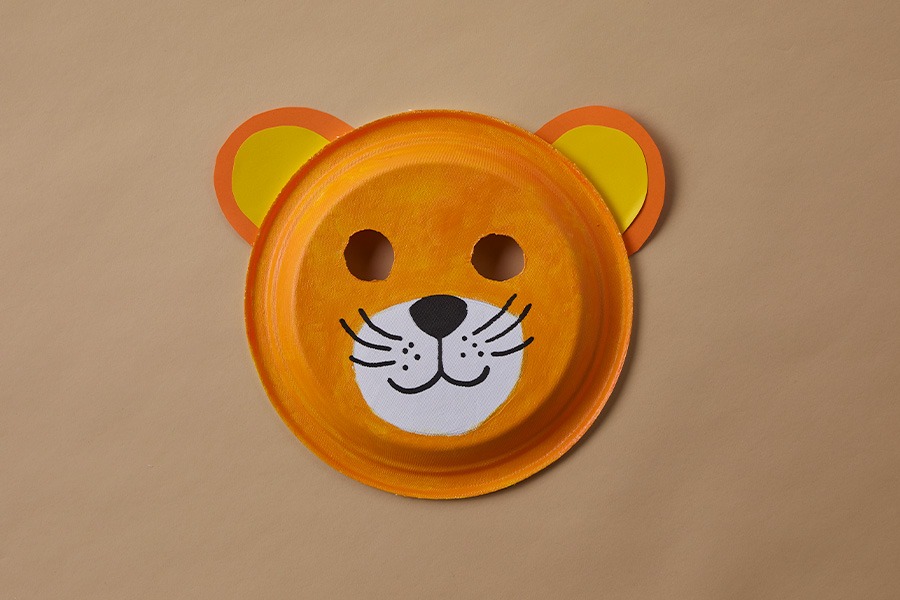 DIY lion mask made from a painted paper plate with cut-out eye holes, paper ears and a drawn face on a plain brown background.