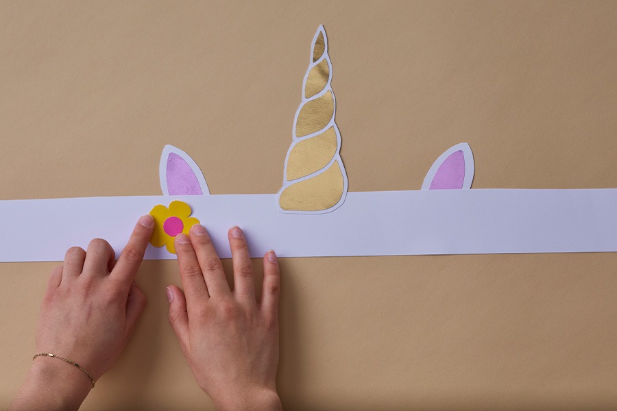 Hands adding a paper flower to a DIY unicorn headband with a gold horn and pink ears on a plain brown background.
