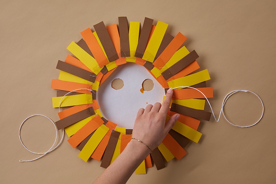 Hand placing a piece of elastic onto a DIY lion mask made from a white paper plate with yellow, orange and brown mane pieces on a brown background.