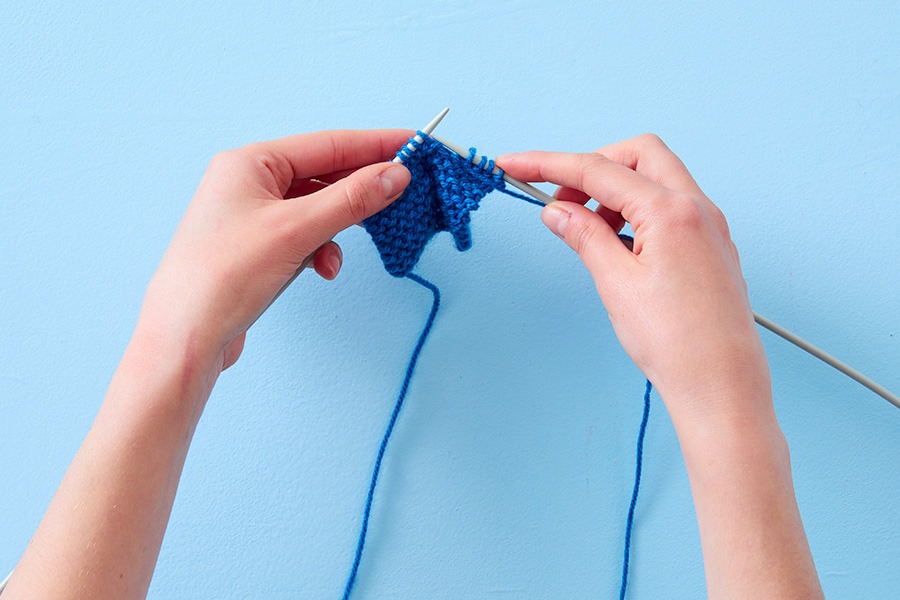 Hands knitting a small piece of blue yarn with knitting needles against a light blue background.