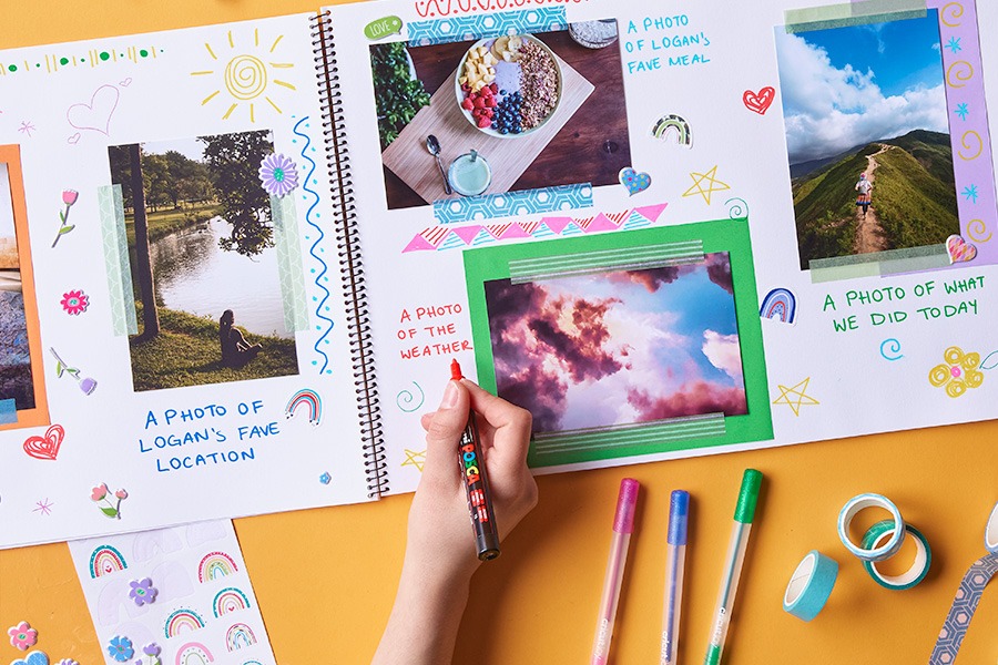 A  hand writing in a scrapbook with photos, stickers and colourful captions in an open spiral album on an orange surface, with pens and washi tape nearby.‍