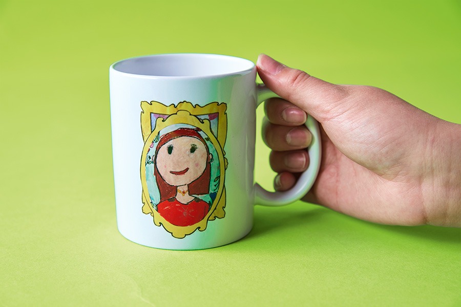 A white mug held in a hand, printed with a child’s colourful portrait drawing on a bright green background.