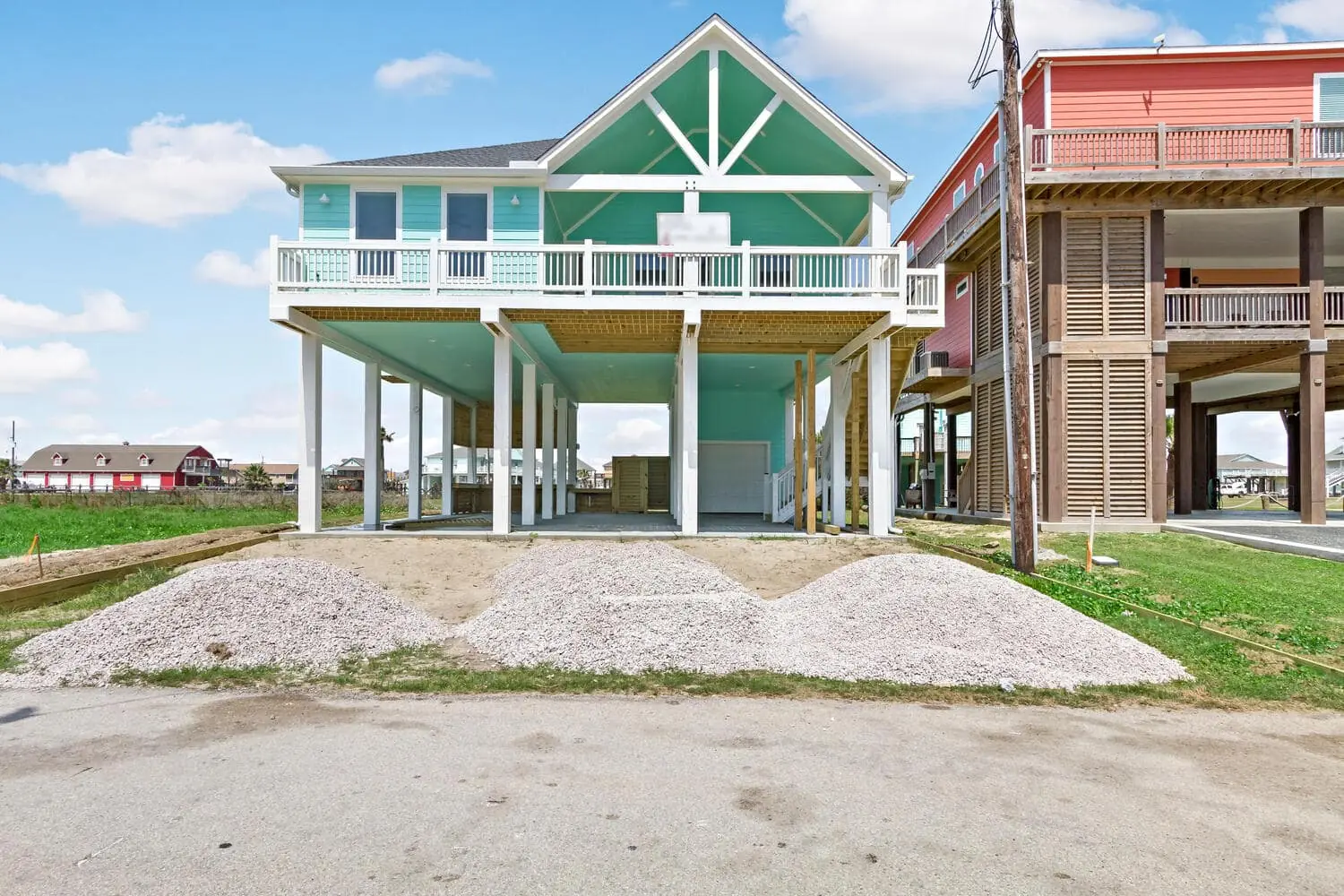 Elevated beach house with a turquoise exterior and white rails above three gravel piles.