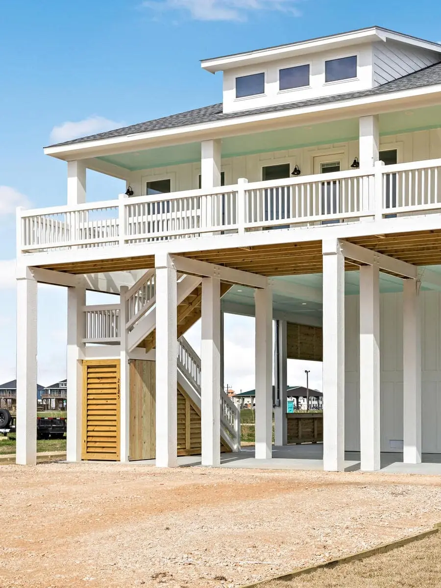 Modern elevated white house with wraparound porch and exterior staircase on gravel lot.