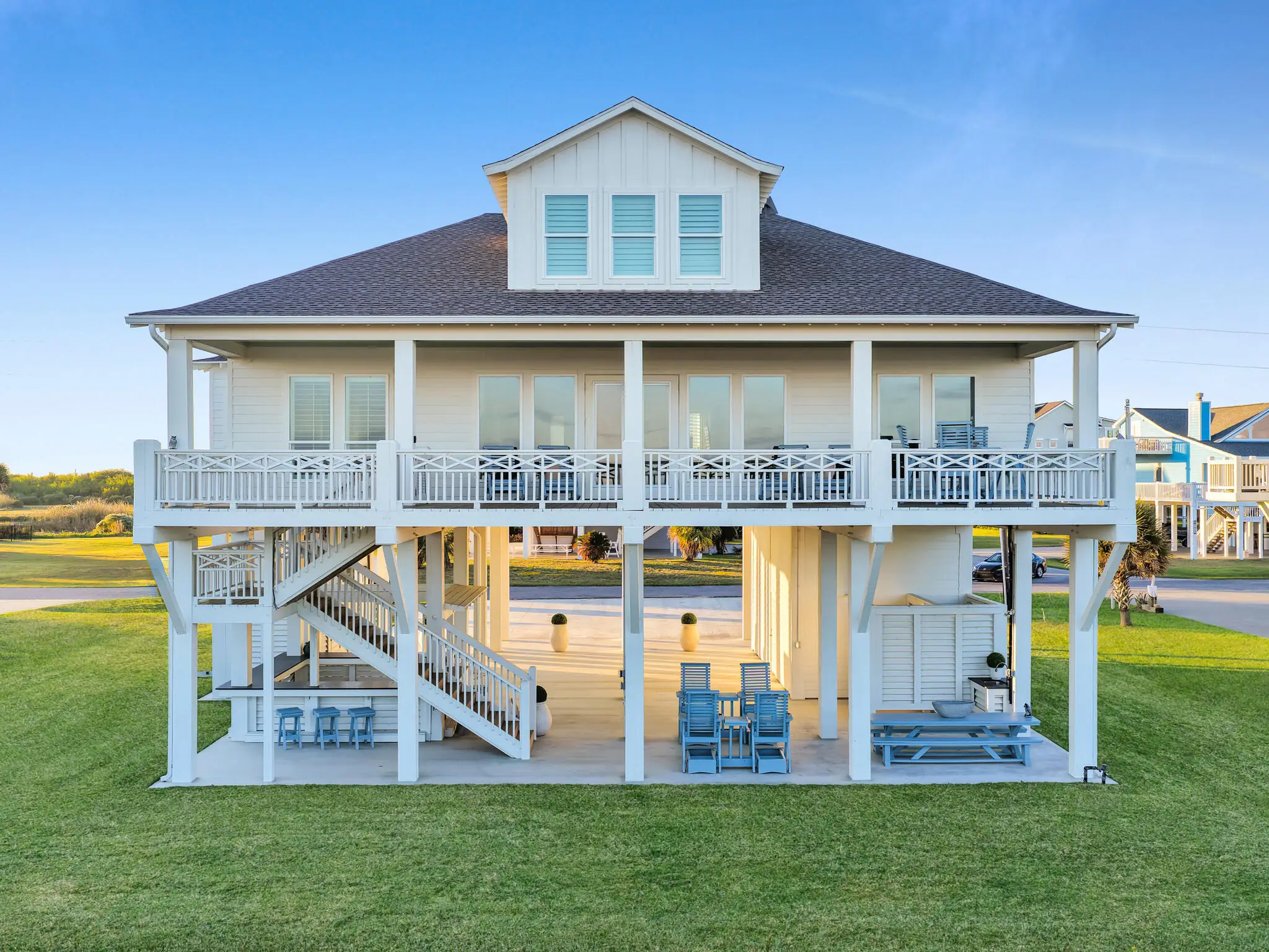 Elevated white house with a large porch, blue outdoor furniture, and stairs leading upstairs.