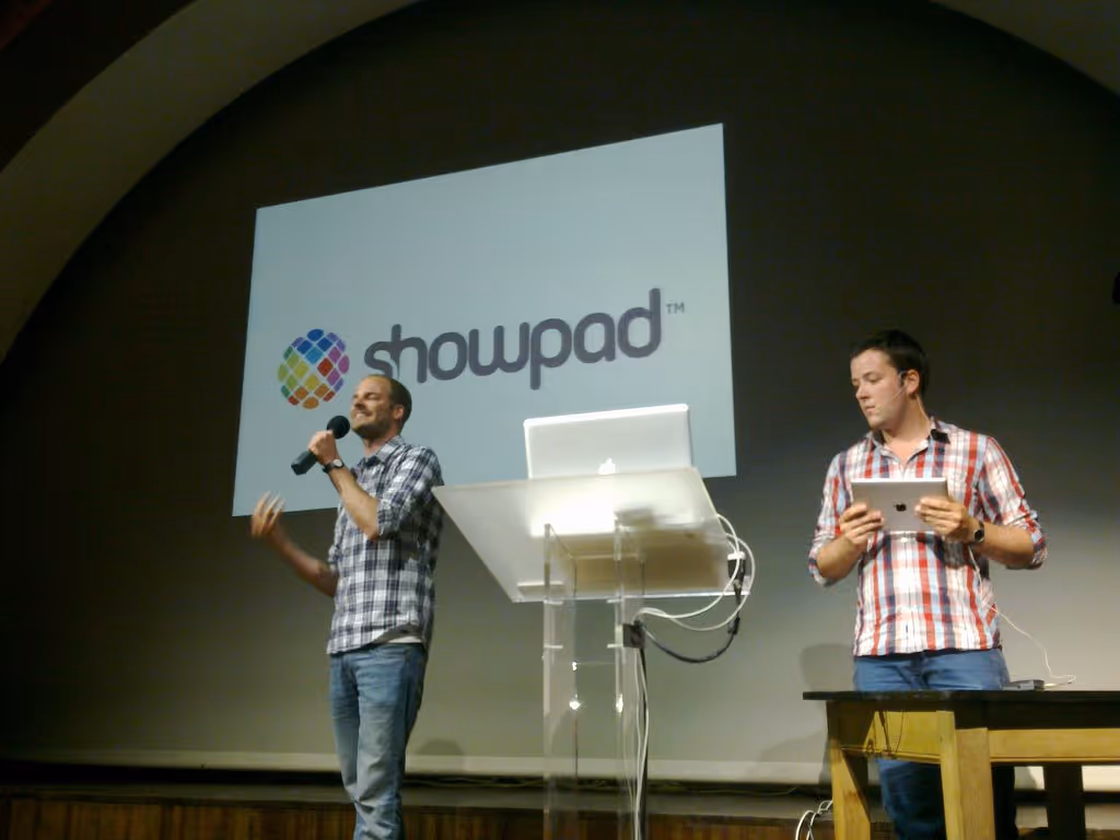 Two men in casual plaid shirts giving a presentation on stage with a Showpad logo displayed on a large screen behind them.