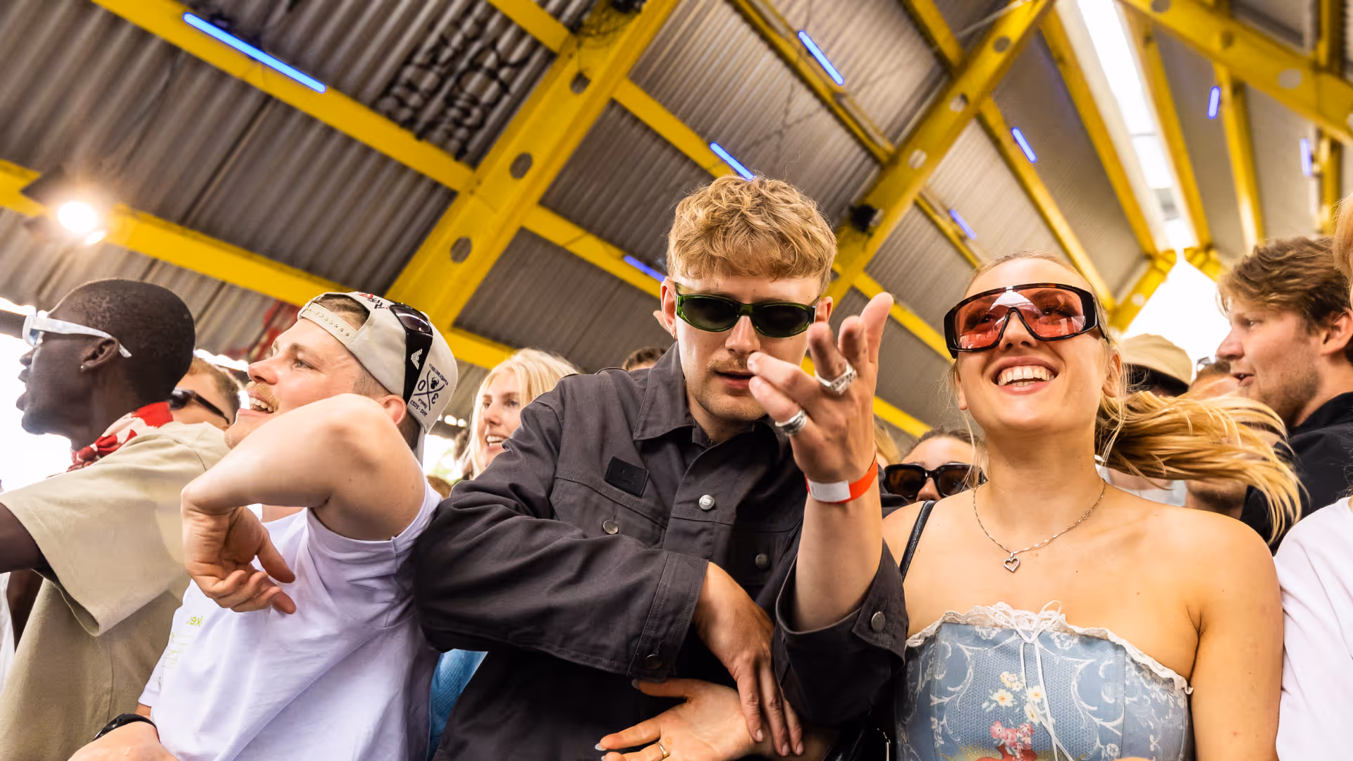 A group of young people wearing sunglasses enjoying a lively event under a yellow-beamed roof.
