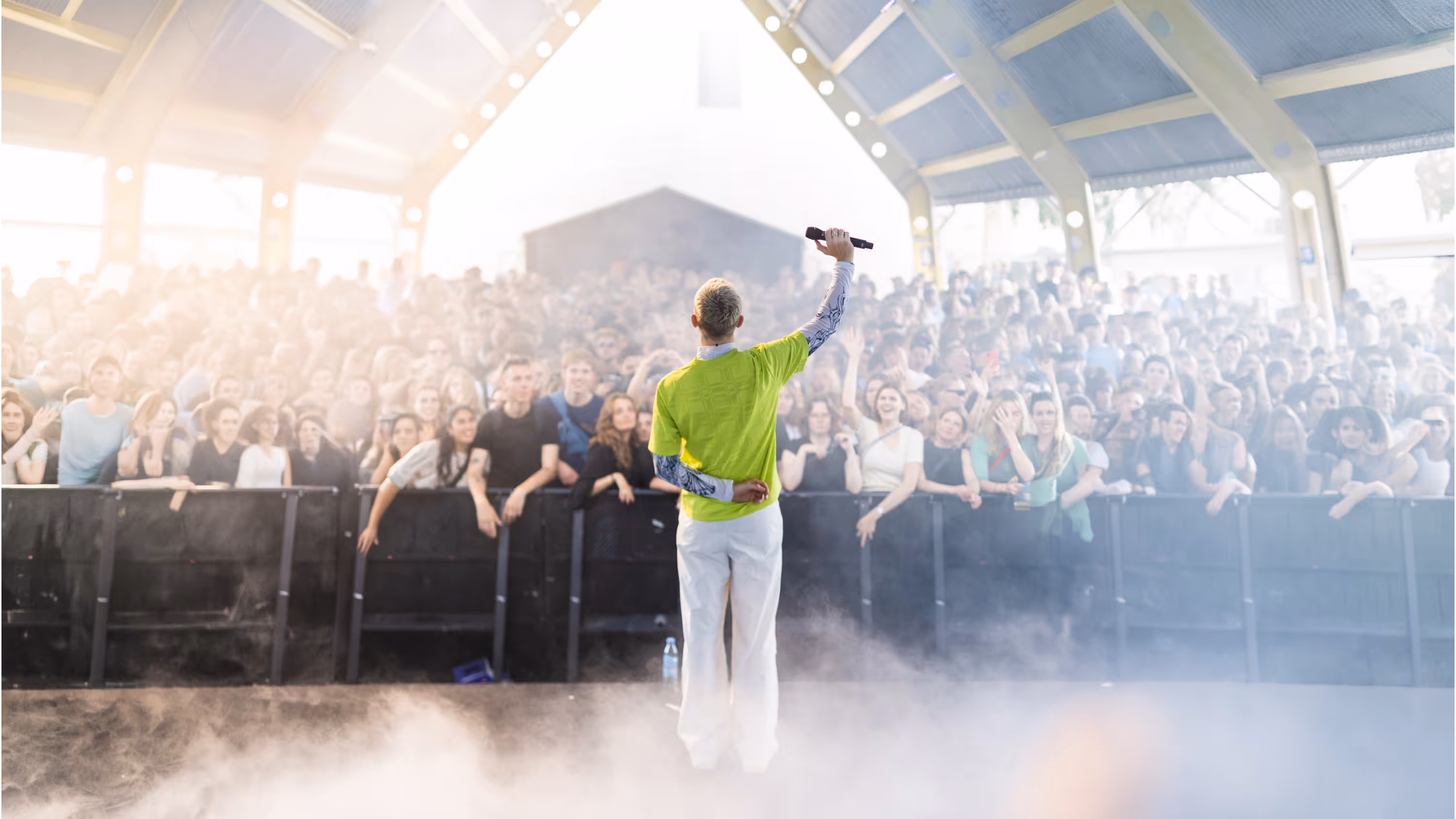 Performer in a lime green shirt and white pants holding a microphone facing a large crowd under a covered venue.