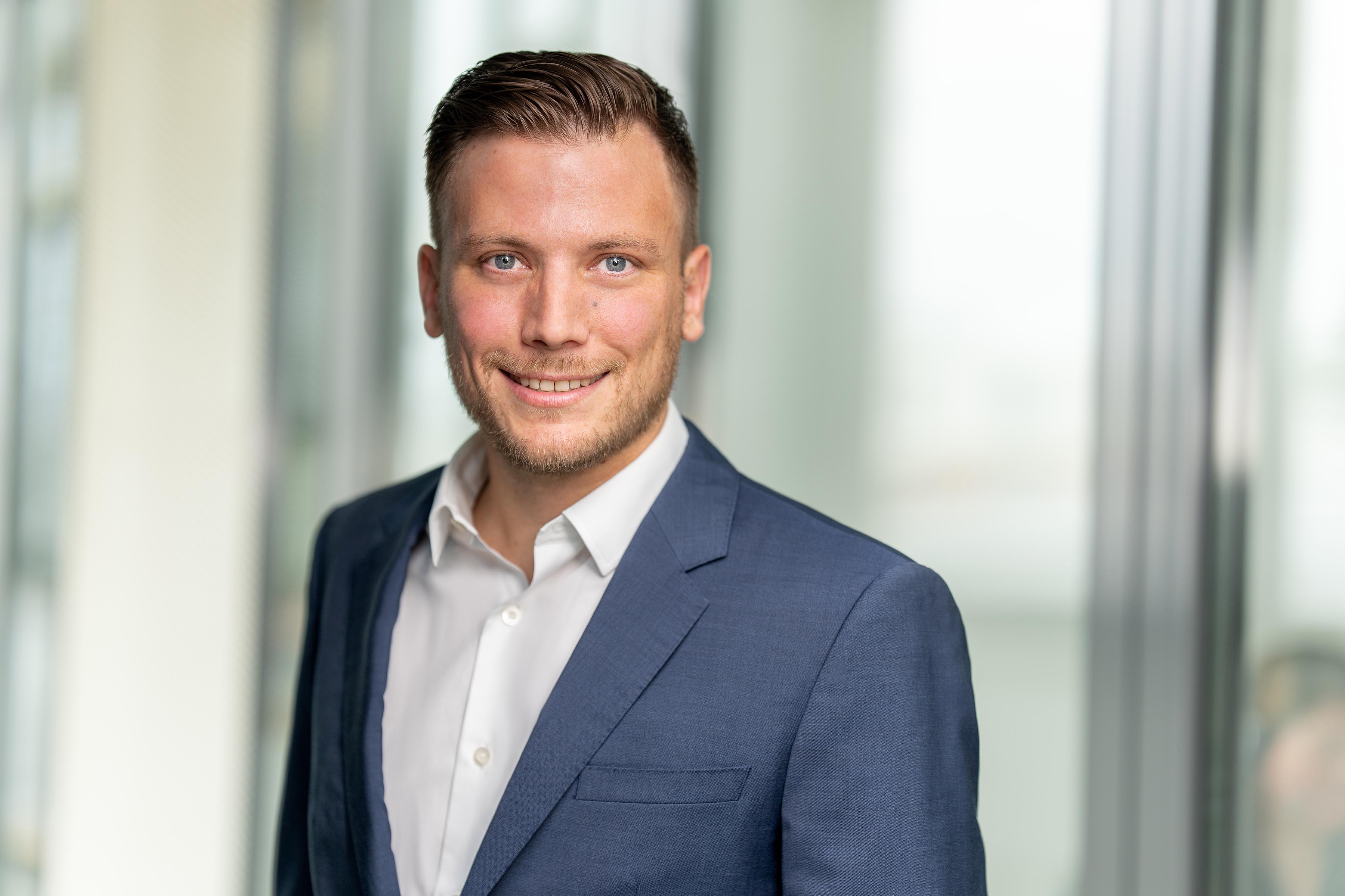 Young man with a short beard in a blue suit and white shirt in a glazed office - Vielen Dank team portrait.