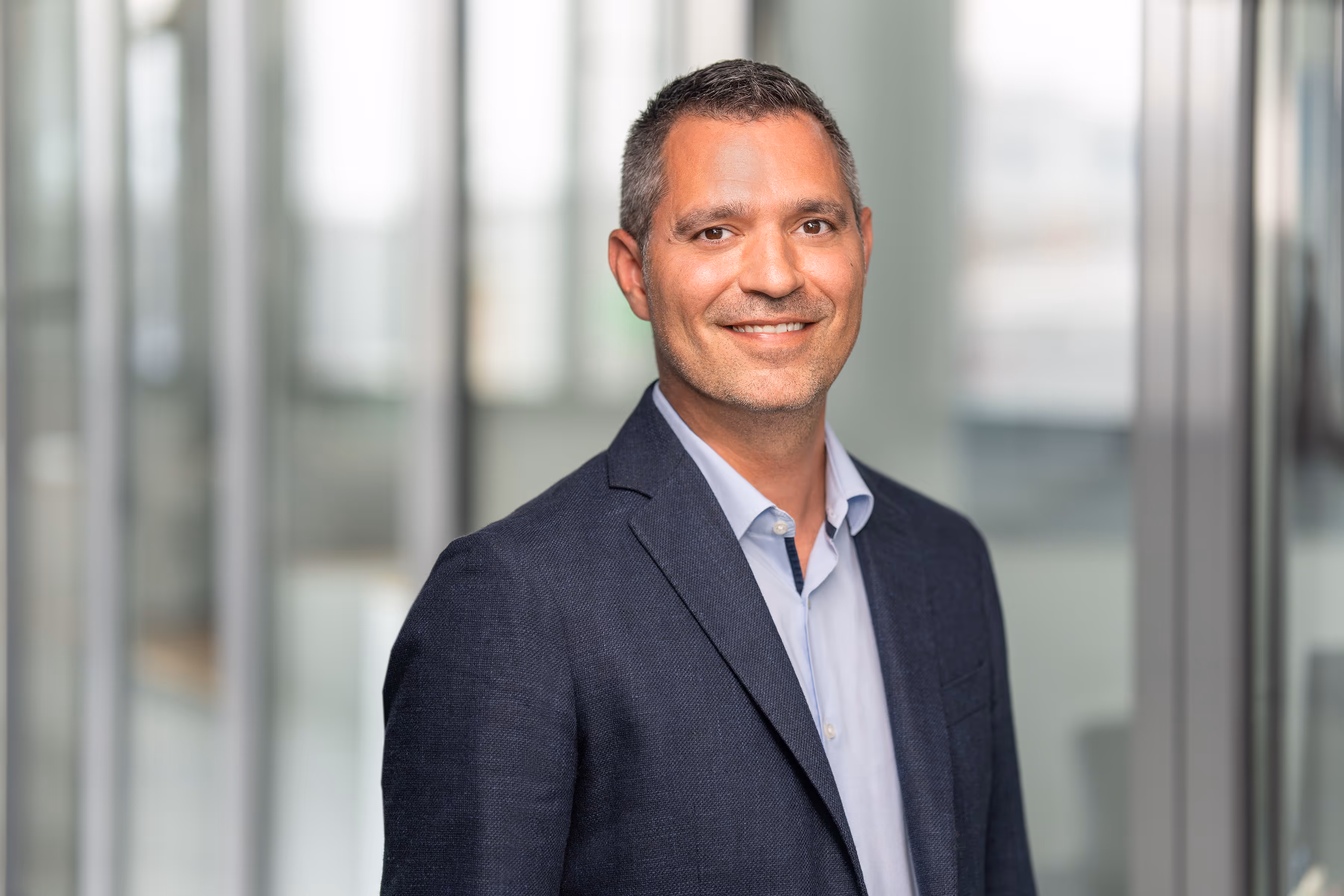 Man in a navy blazer and open-collar shirt in a glazed office corridor - Vielen Dank team portrait.
