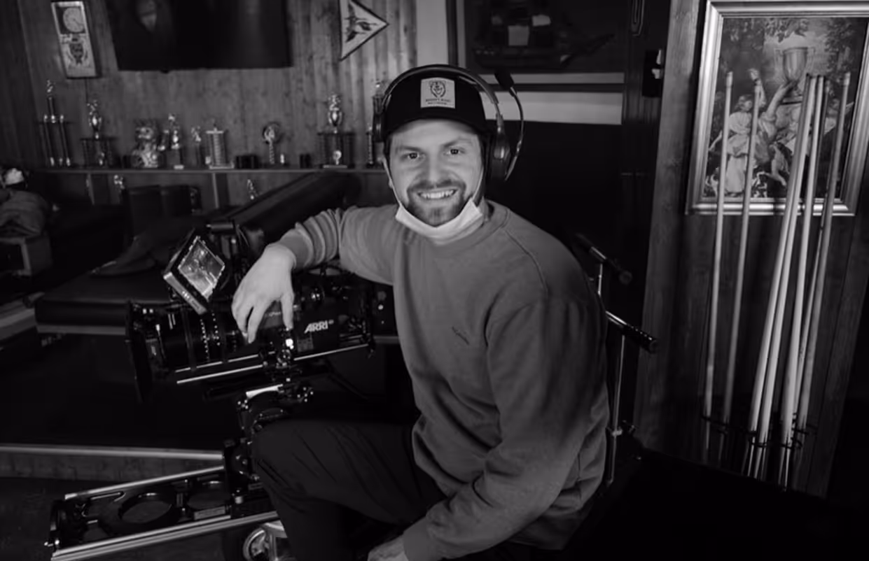Man wearing headphones and a cap sitting behind a professional ARRI camera in a room with trophies and pool cues.