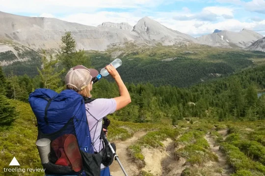 A lady drinking from a water filter while looking at a mountain.