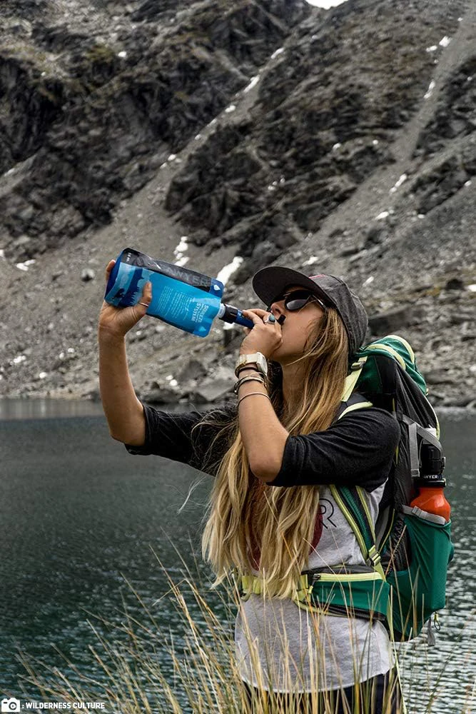 A woman drinking from a Sawyer mini water filter and pouch by a lake in the mountains