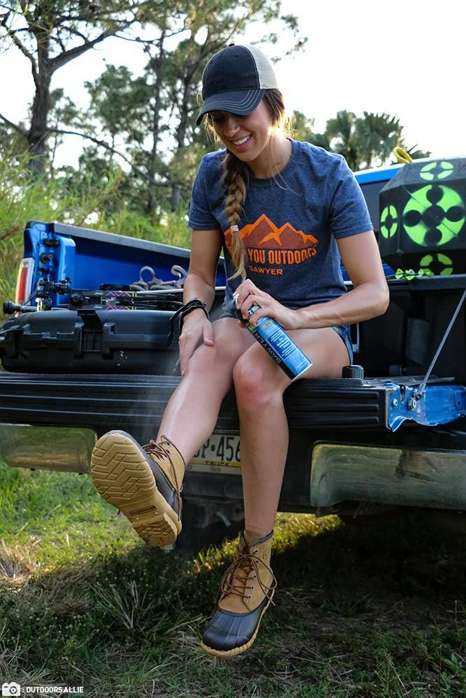 A woman spraying Sawyer picaridin insect repellent on legs sitting on the back of a truck bed