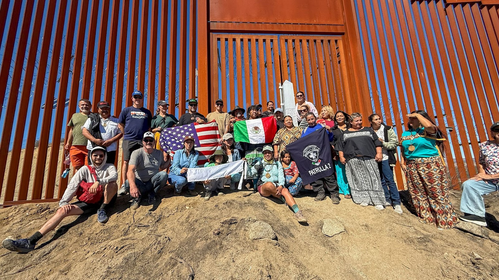 A crowd gathers at the US-Mexico border wall with several flags to celebrate the beginning of the trail