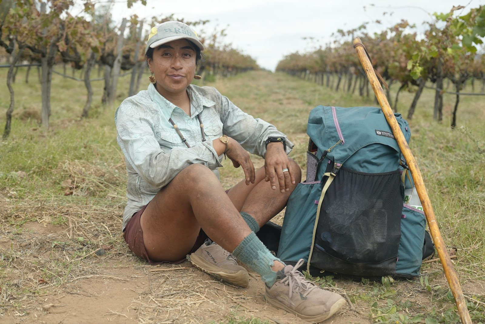 Woman sitting in vineyard, camping gear and trekking pole beside her