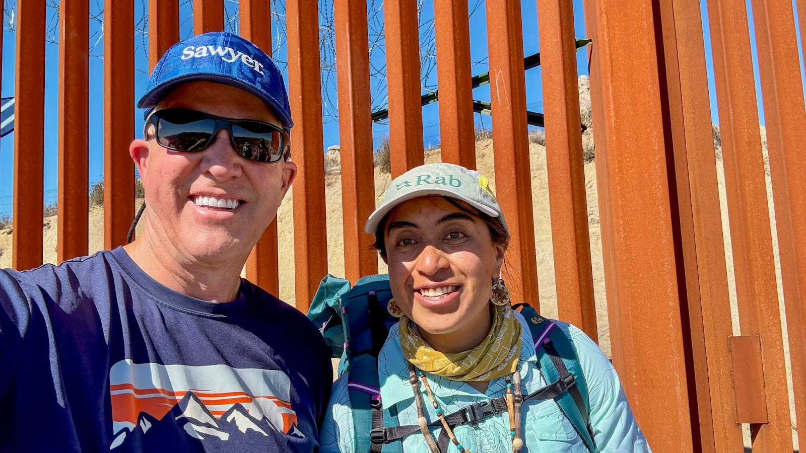 Man and woman smiling with US-Mexico border wall in the background