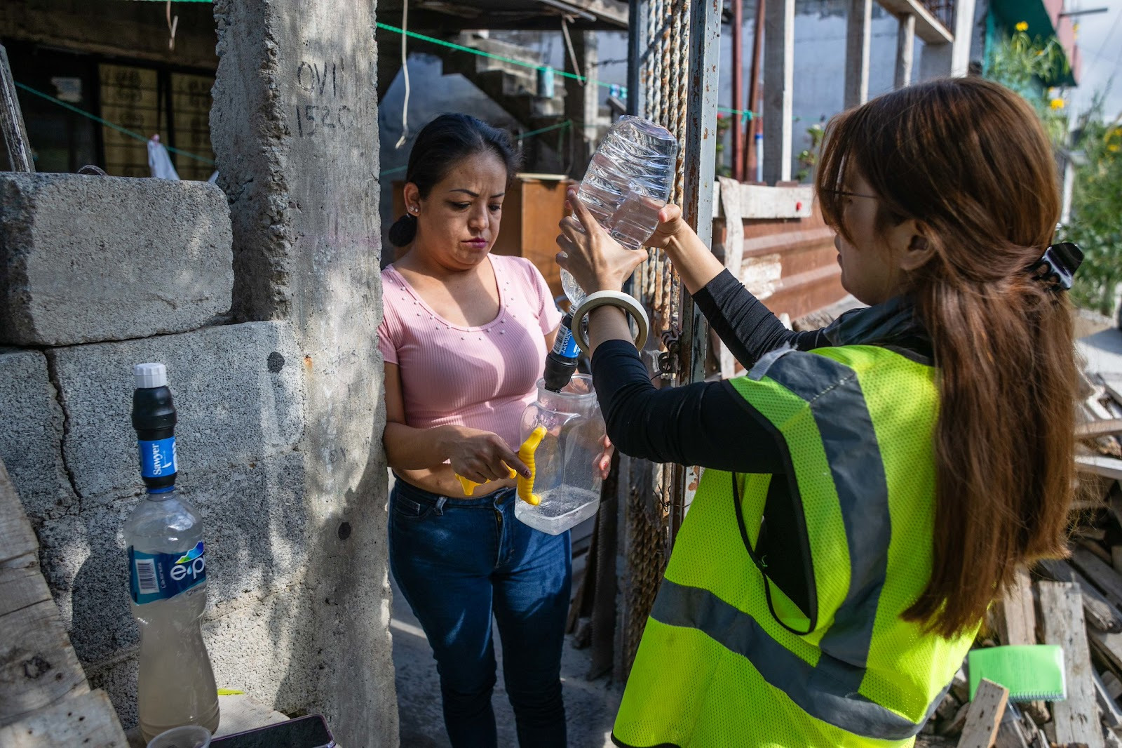 two women standing in the threshold of a home, one in a safety vest squeezing clean water through a bottle into a waiting container held by the other woman