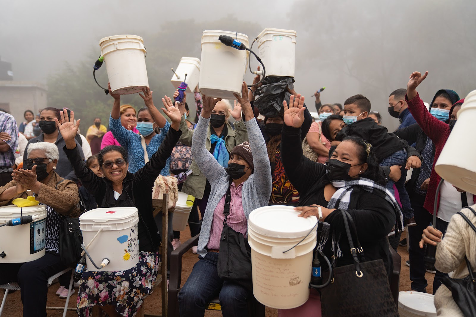 a crowd of women gather on a foggy day and hold their bucket fitration systems above their heads in an expression of joy