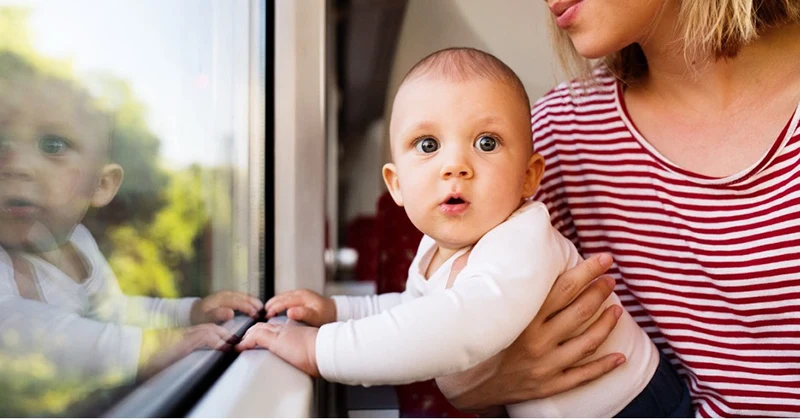 A lady holding a baby by a window.
