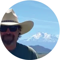 Man wearing sunglasses and a cowboy hat with snow-covered mountains in the background.