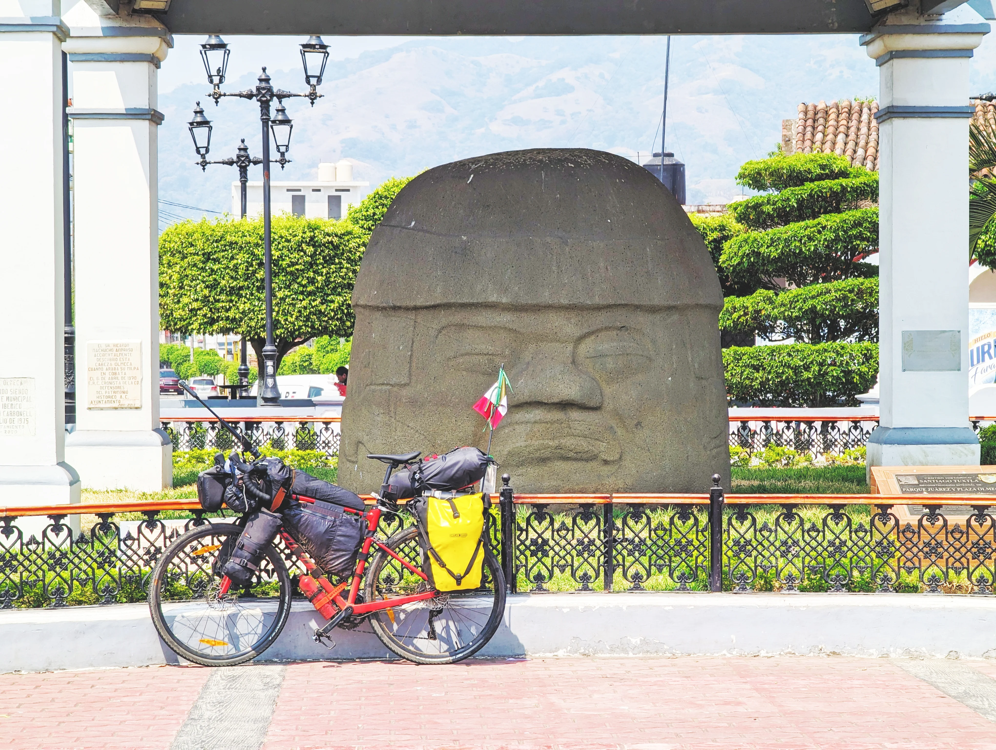 A red bikepacking set up leans against a large stone monument and small decorative fence on a brick street, with a blue sky and green topiaries in the background.