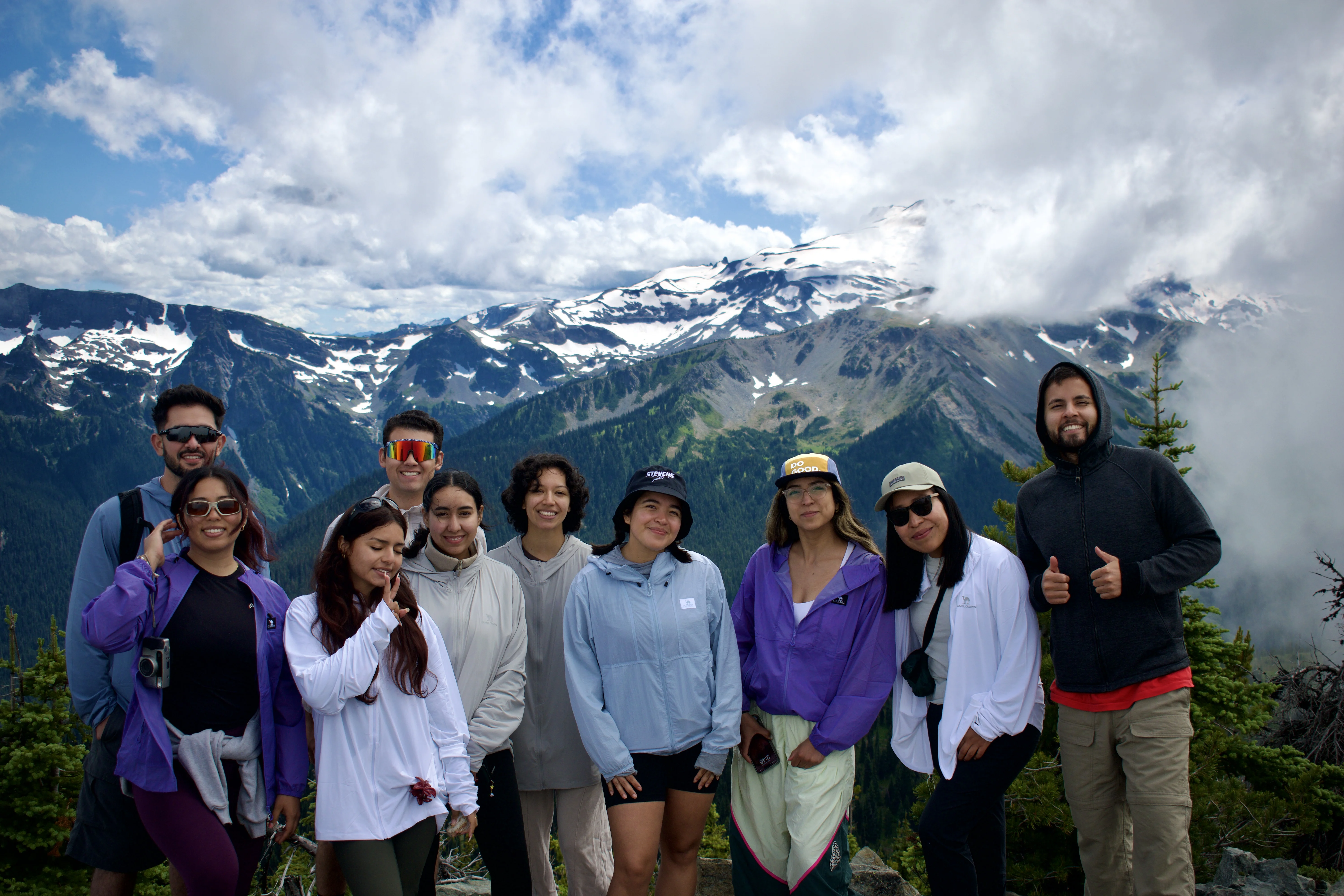 Group of men and women standing facing forward outdoors and smiling for the camera in front of snow capped mountains in background