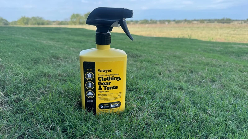 Yellow spray bottle of Sawyer Premium Insect Repellent placed upright on green grass in an open field under a blue sky.