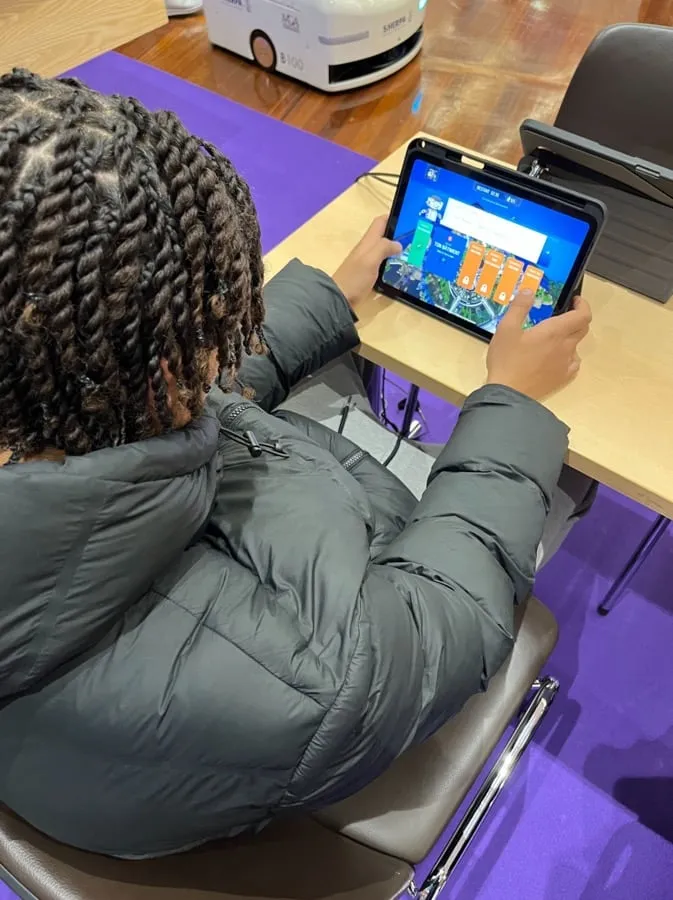 Person with braided hair wearing a dark jacket sitting on a chair and interacting with a tablet on a desk.