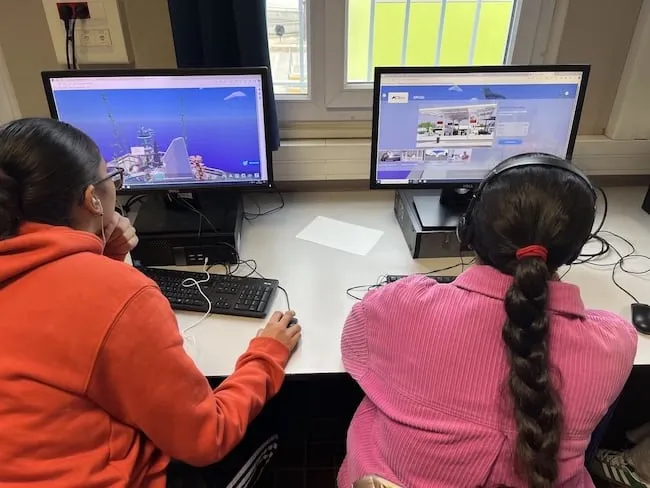 Two people seated side by side at a desk using desktop computers, viewed from behind.