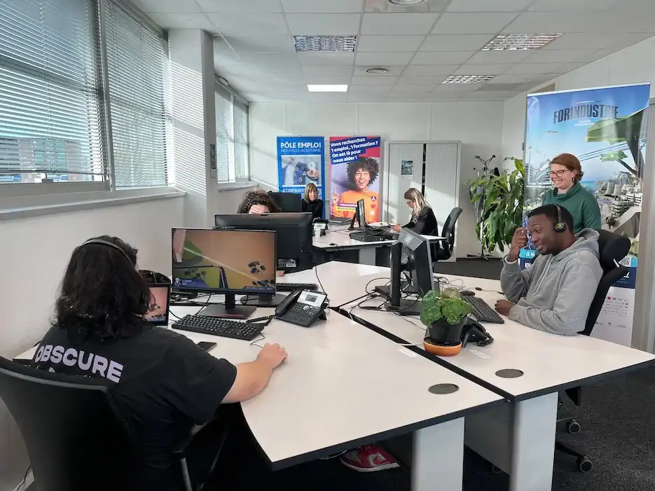 Office with several people working at desks with computers and wearing headsets, a woman standing and smiling.