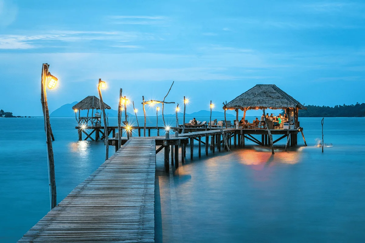Pier with illuminated lanterns leading to a thatched-roof hut restaurant over calm sea at dusk.