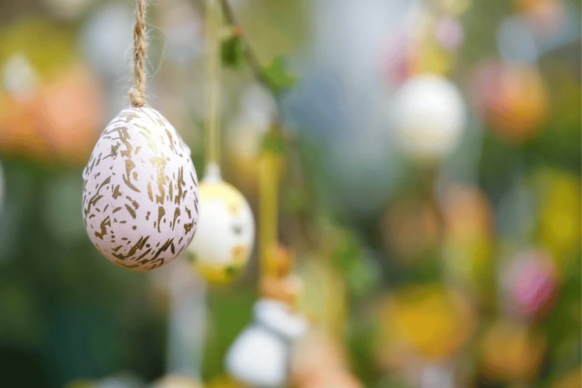 A decorative pink Easter egg with gold patterns hangs on a tree, surrounded by softly blurred colorful eggs and greenery.