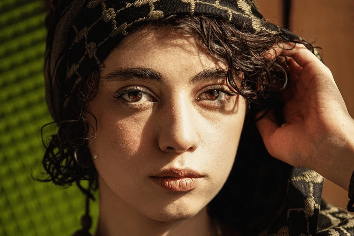 A close-up view of a person wearing a patterned scarf, with a hand adjusting their curly hair, and vibrant green textured background.