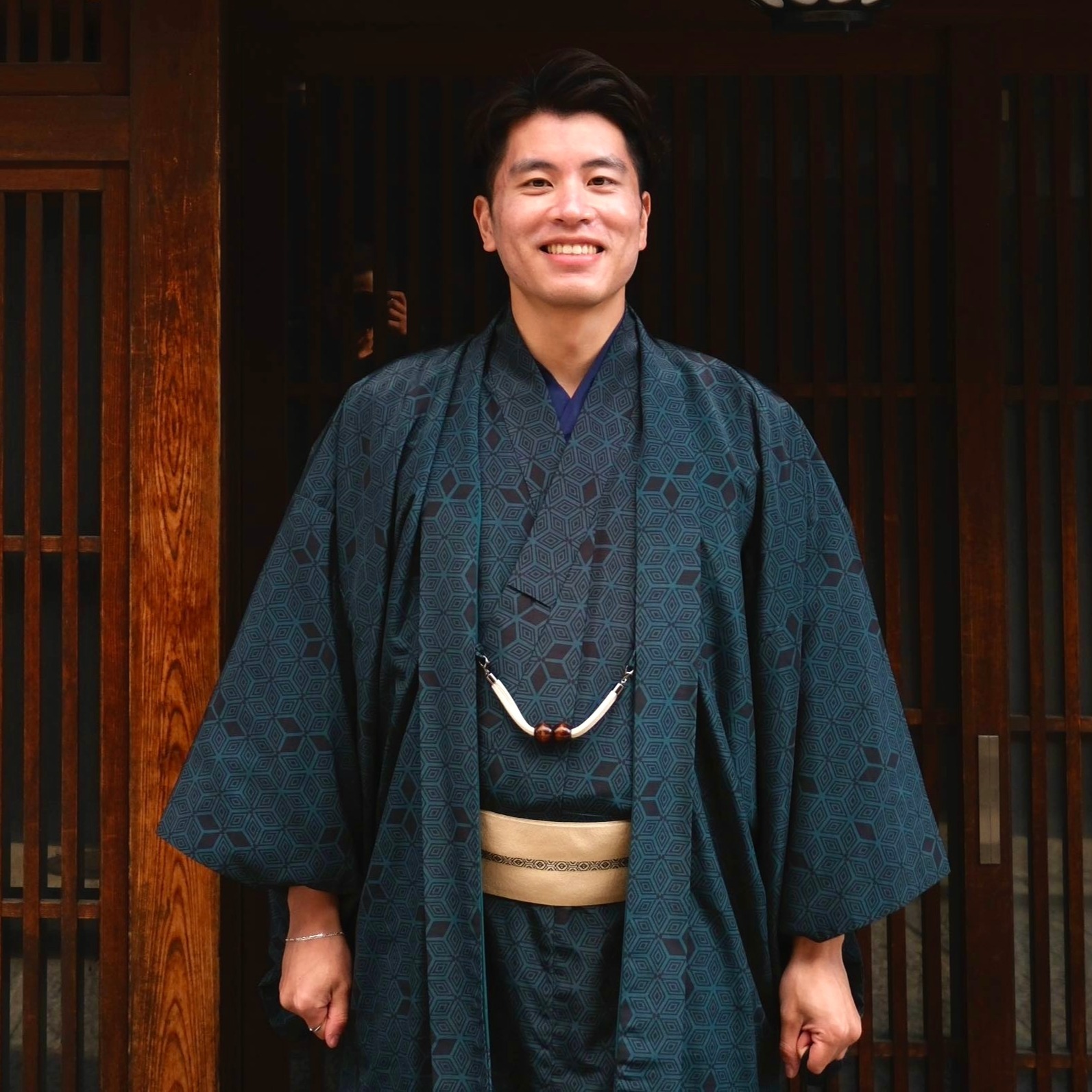 Smiling man wearing a dark patterned traditional Japanese kimono standing in front of wooden sliding doors.