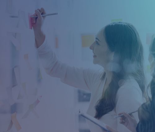 Woman in pink sweater writing on sticky notes during a collaborative planning session, smiling as she posts a red note to a glass board filled with colorful notes.