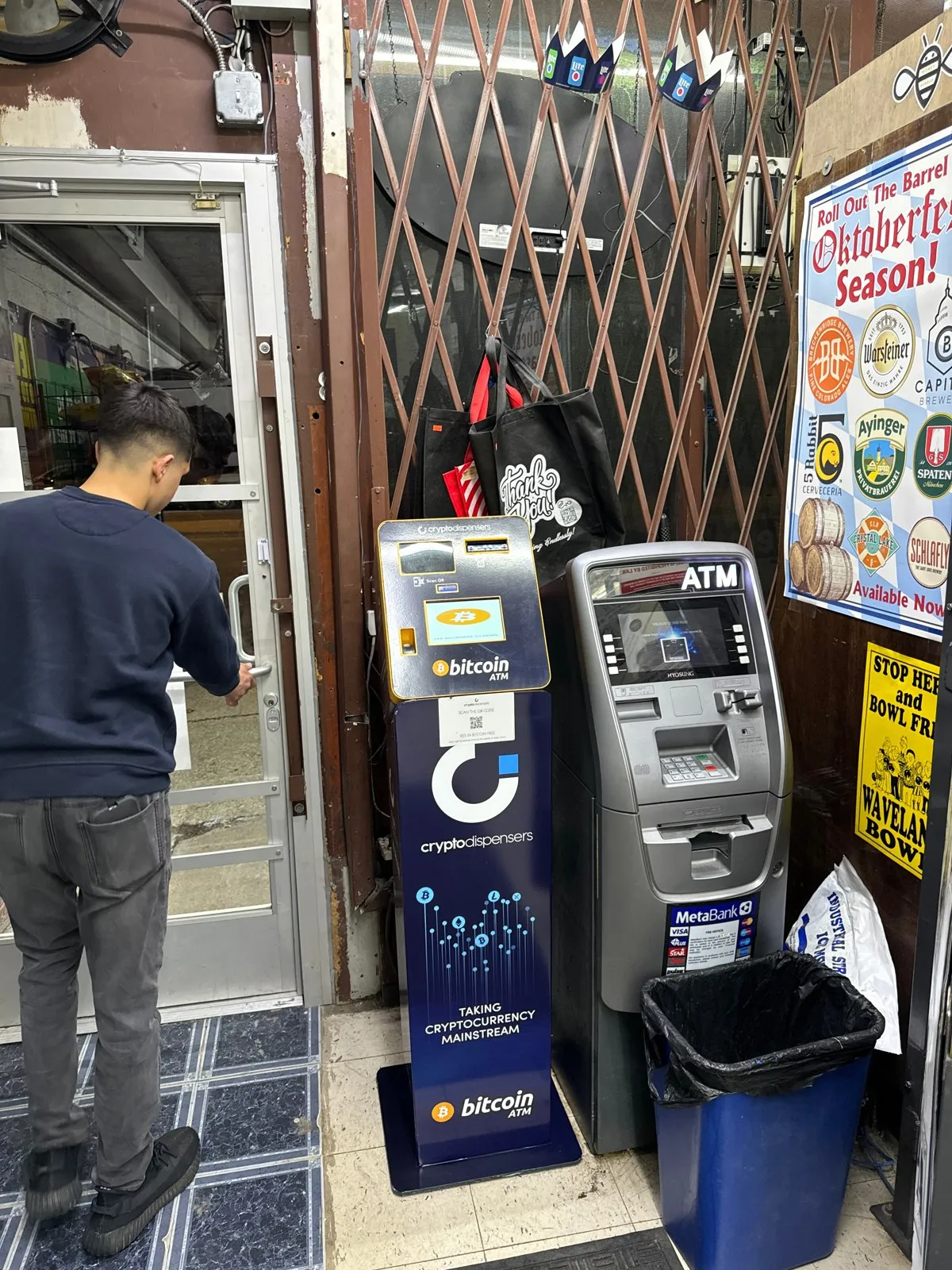 A blue Bitcoin ATM and a gray cash machine standing near a storefront security gate.