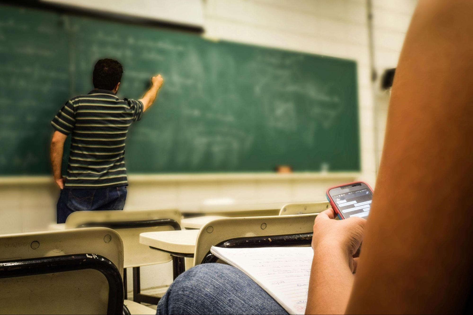 Teacher writing on a chalkboard while a student looks at their phone during class
