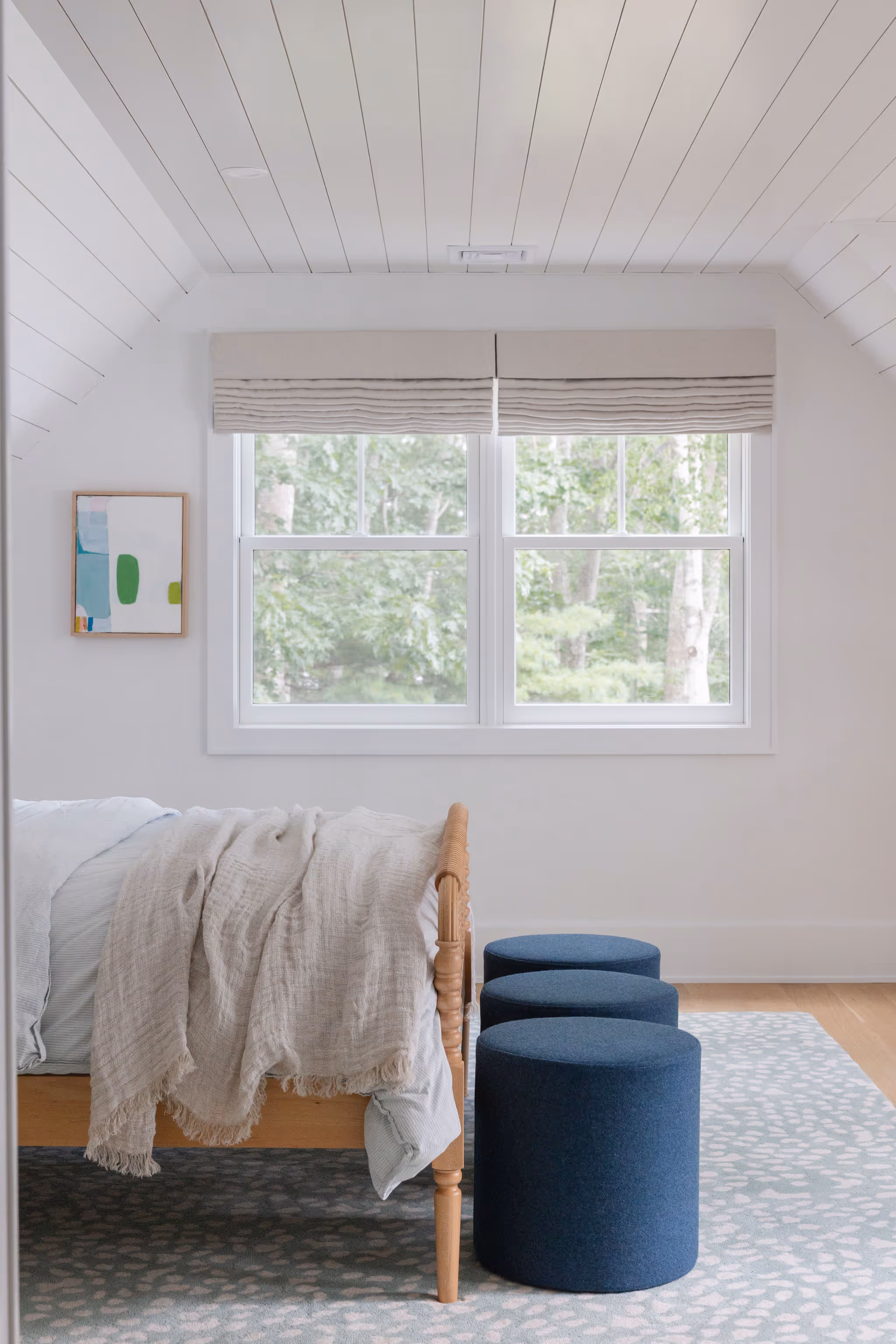 Minimalist bedroom with wooden bed, blue poufs, and windows overlooking trees