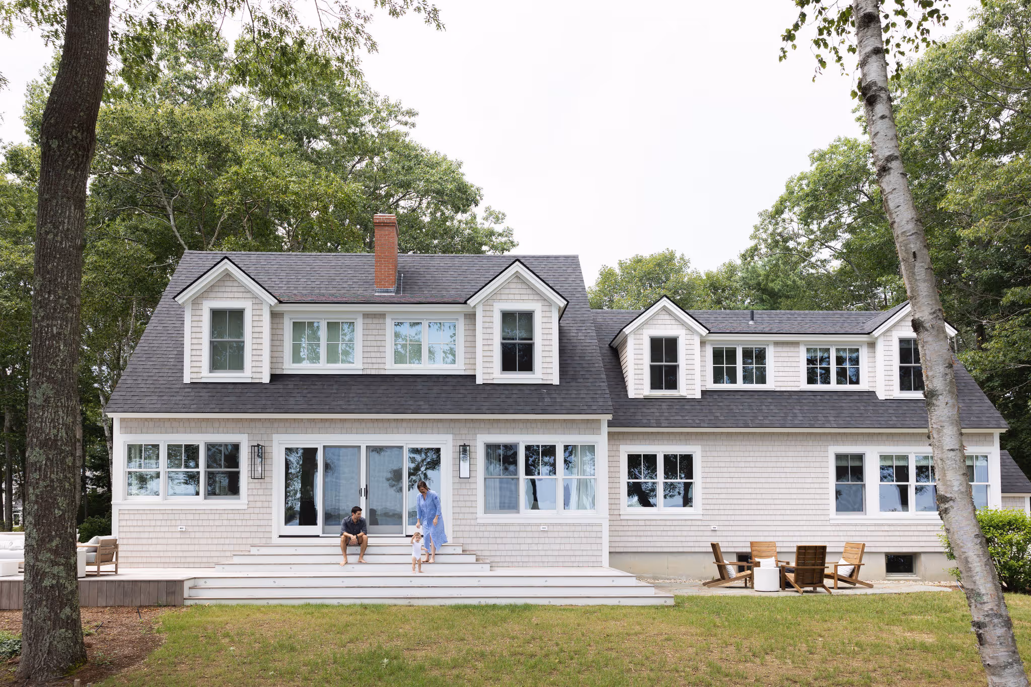 White coastal home with two people sitting on front porch, surrounded by trees