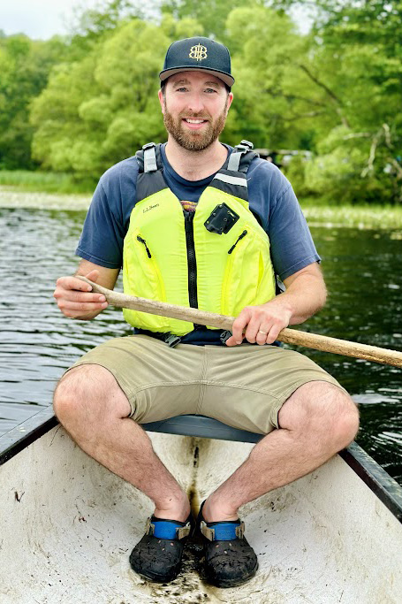 Person sitting in boat holding rower on body of water with trees in background