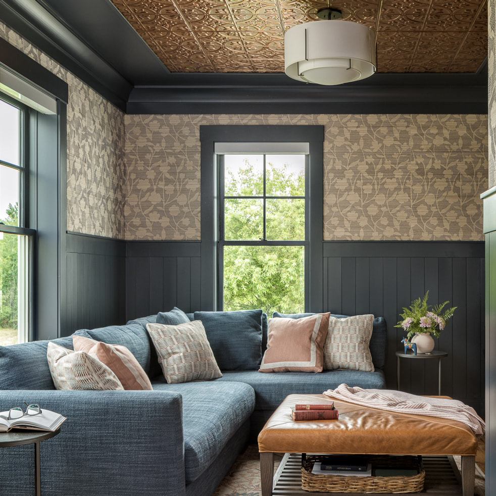 Cozy living room with a large blue sectional sofa adorned with patterned and solid pillows, a brown leather ottoman with books and a throw, floral wallpaper above dark wood paneling, a window showing outdoor greenery, and a modern ceiling light fixture on a patterned ceiling.