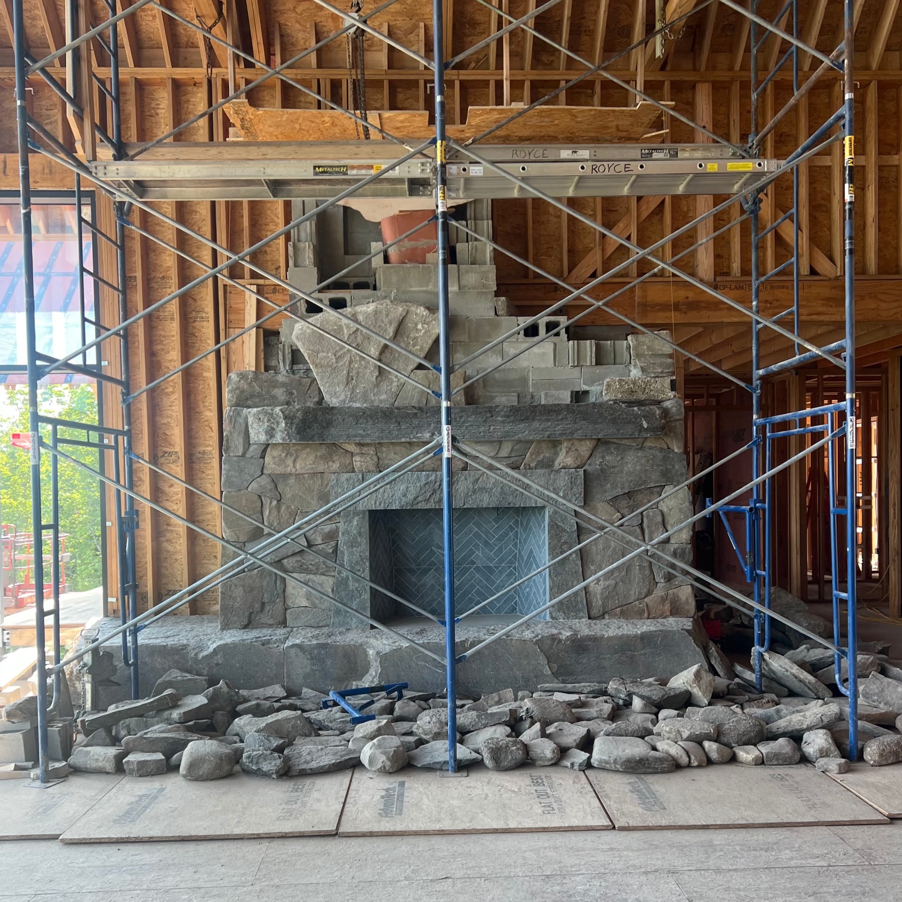 Large stone fireplace under construction inside a wooden frame building, surrounded by metal scaffolding and loose stones on the floor in front.