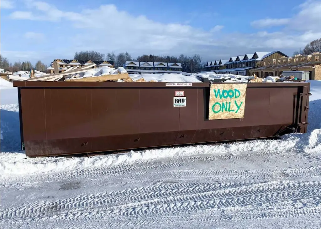Large brown dumpster labeled 'WOOD ONLY' filled with wood scraps and covered in snow at a snowy construction site.