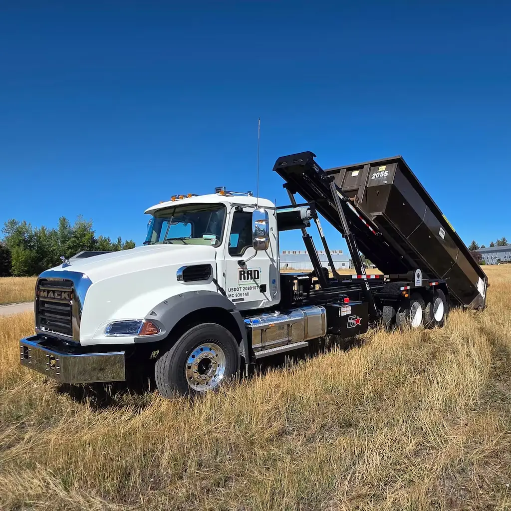White Mack roll-off truck with raised black container in a grassy field under clear blue sky.