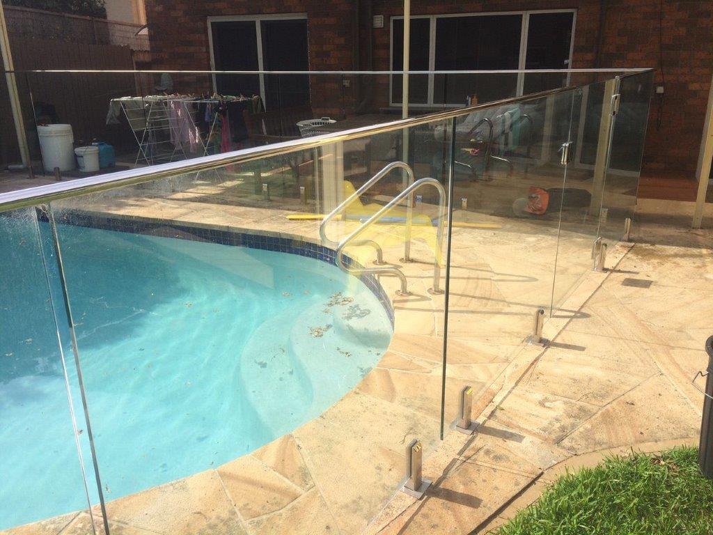 Curved swimming pool with metal handrails behind a glass safety fence on a stone patio near grass.